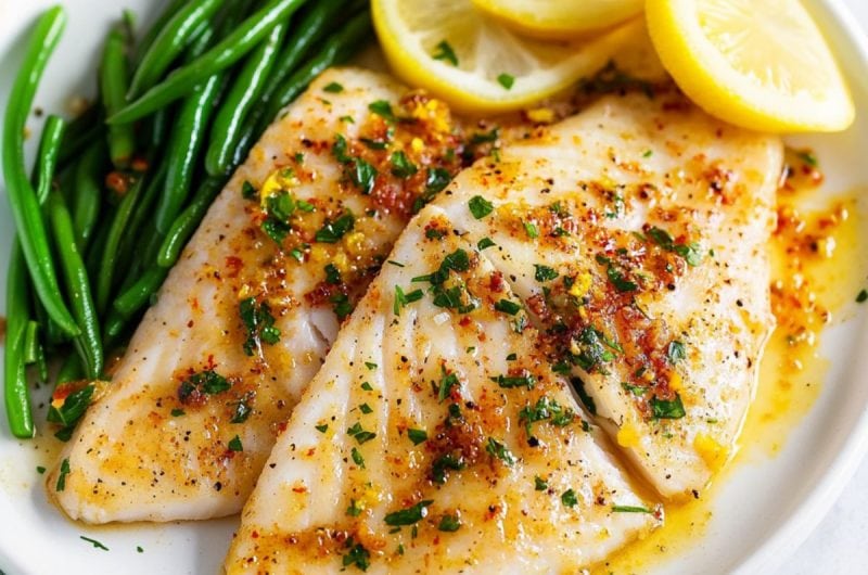 Oven baked tilapia fillet served in a white plate with lemon wedge and steamed beans.