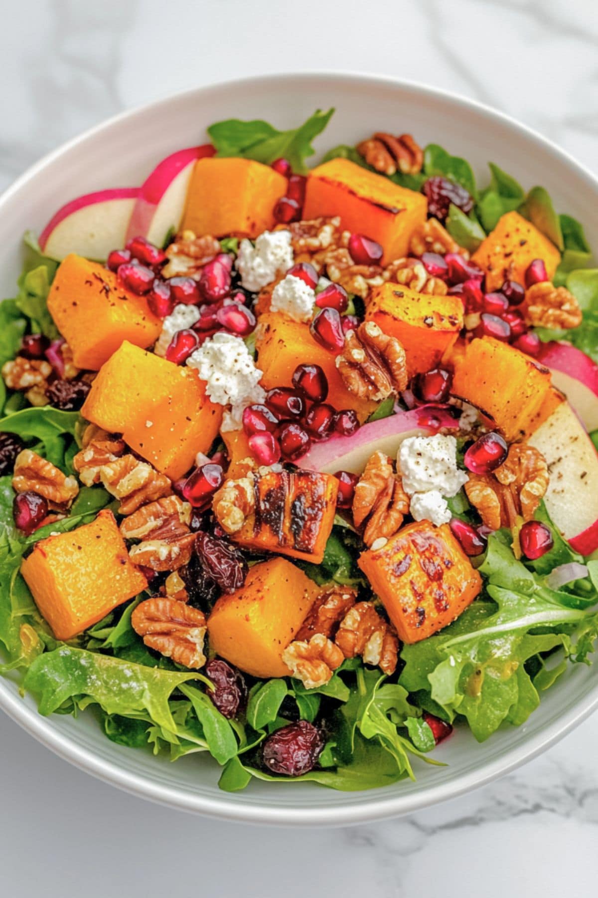 White bowl with a serving of roasted squash, sliced apples, red onion, pomegranate, and goat cheese