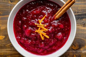 A jar of homemade cranberry sauce with a deep red color, garnished with orange zest and cinnamon.