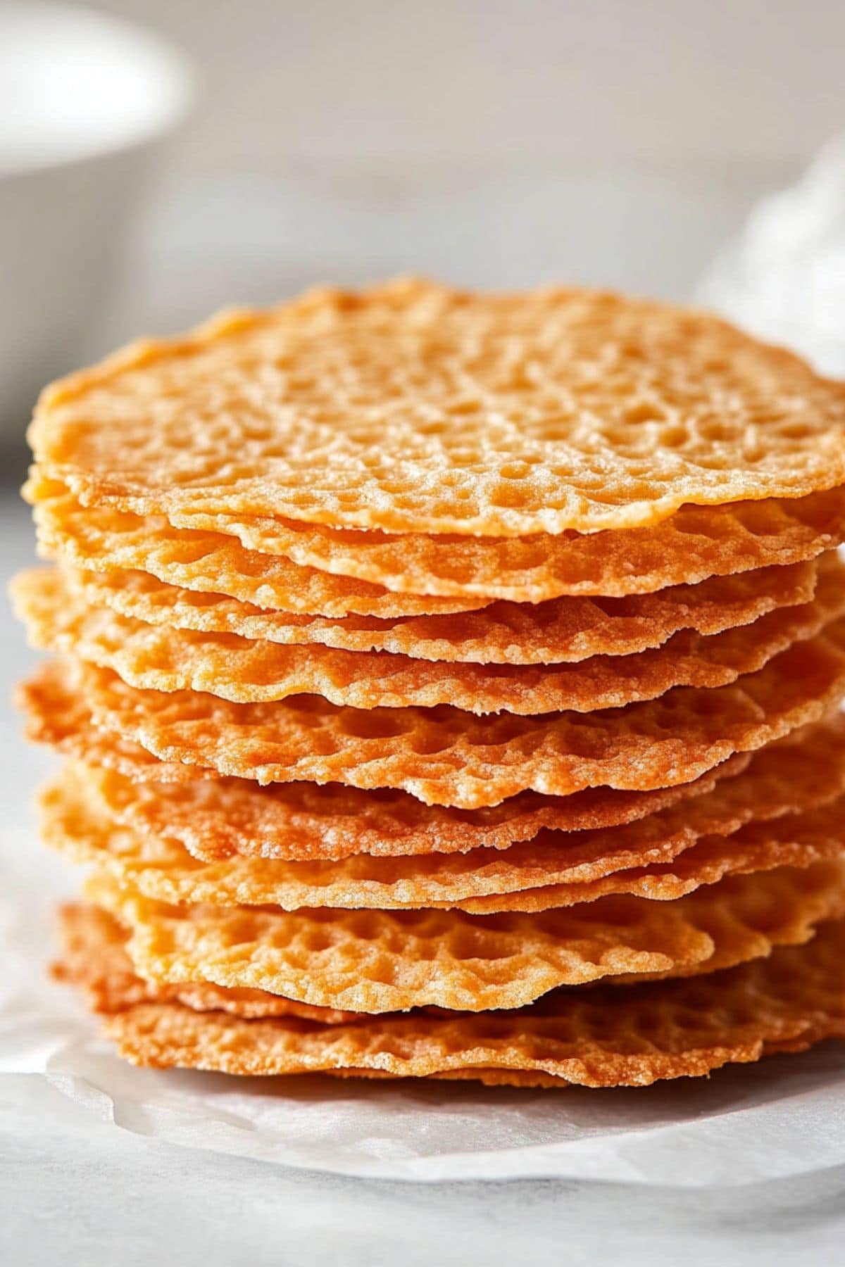 Thin lace like biscuits stacked on top of each other sitting on a white parchment paper.