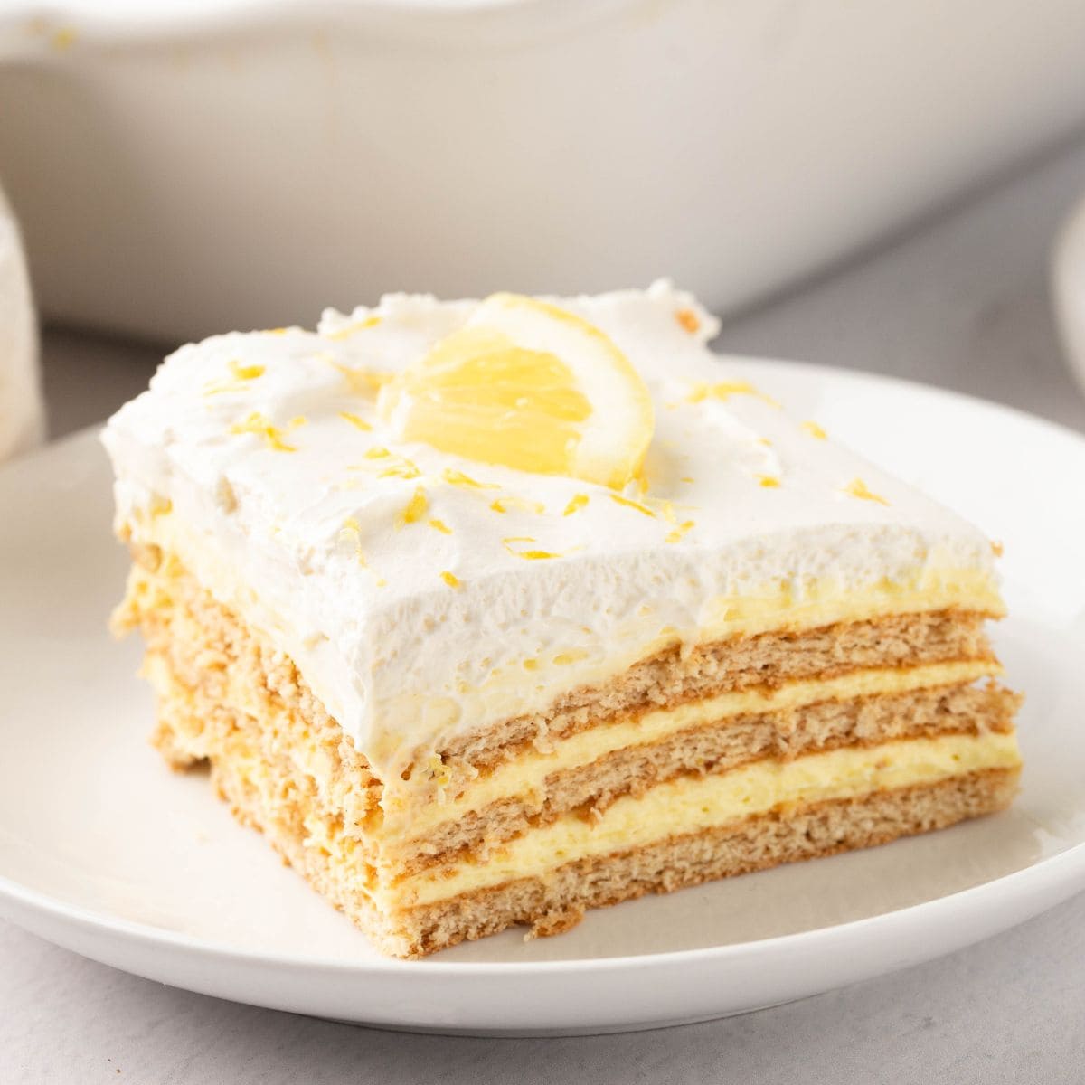 Square slice of lemon icebox cake in a white plate garnished with a lemon slice on top.