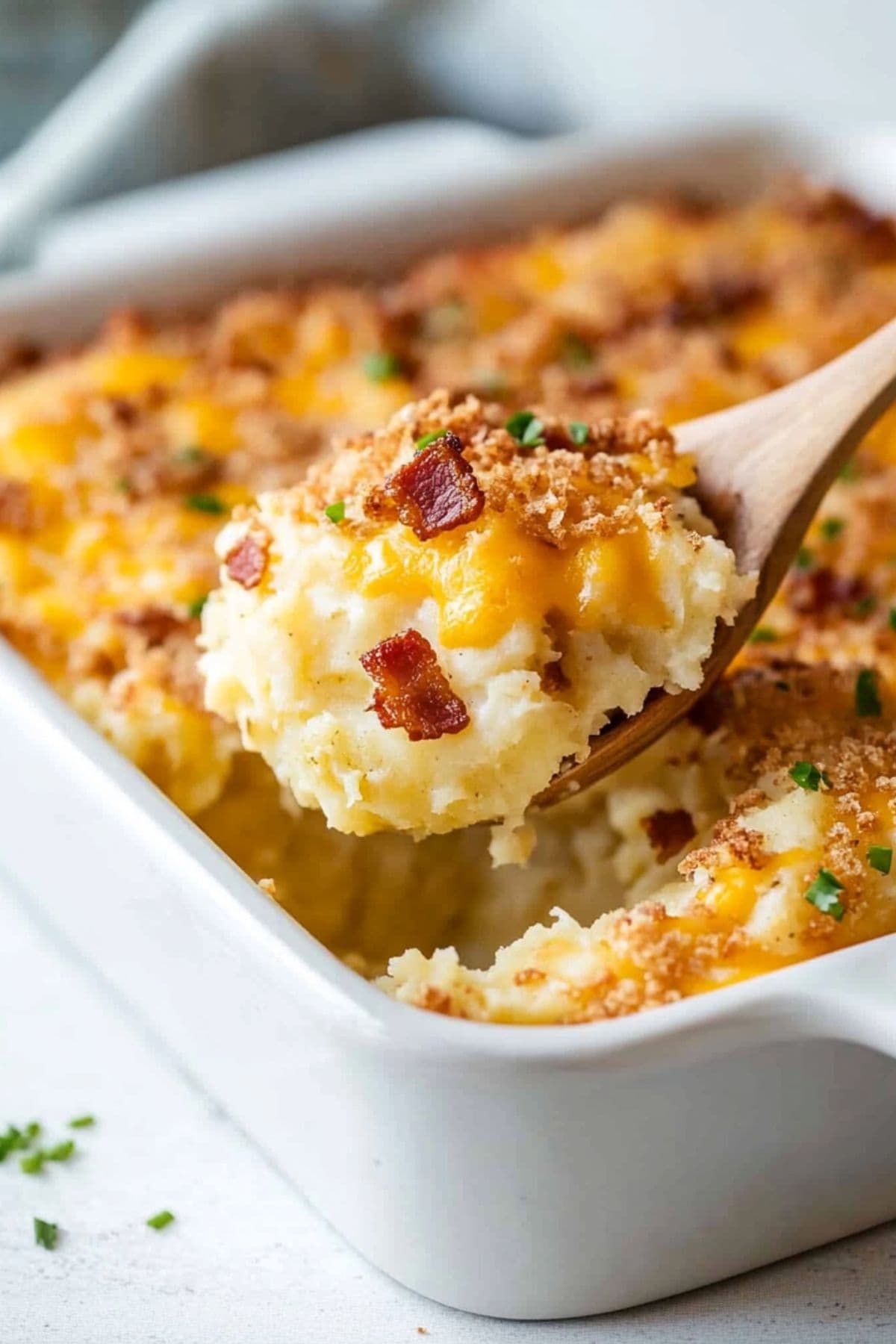 Wooden spoon lifting a serving of cheesy  mashed potato casserole from baking dish. 