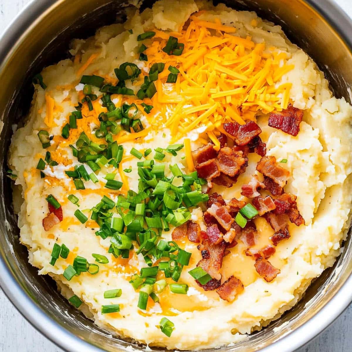 Mashed potatoes with crumbled bacon, cheddar cheese and green onions in a large metal mixing bowl.