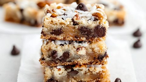 Square magic cookie bars on a parchment paper, with visible chocolate chips, coconut shavings, and a golden graham cracker base.