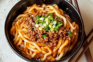 Udon noodles with ground beef and savory sauce served in a black bowl with chopped onions and sesame seeds garnish on top.