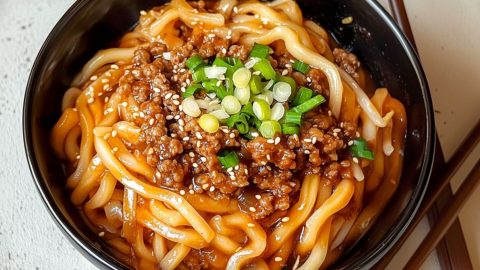 Udon noodles with ground beef and savory sauce served in a black bowl with chopped onions and sesame seeds garnish on top.