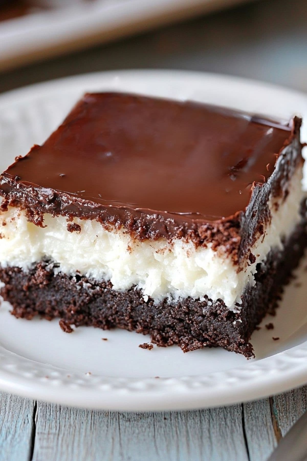 A square slice of chocolate brownie with creamy coconut filling topped with chocolate ganache served on a white sauce plate, close up