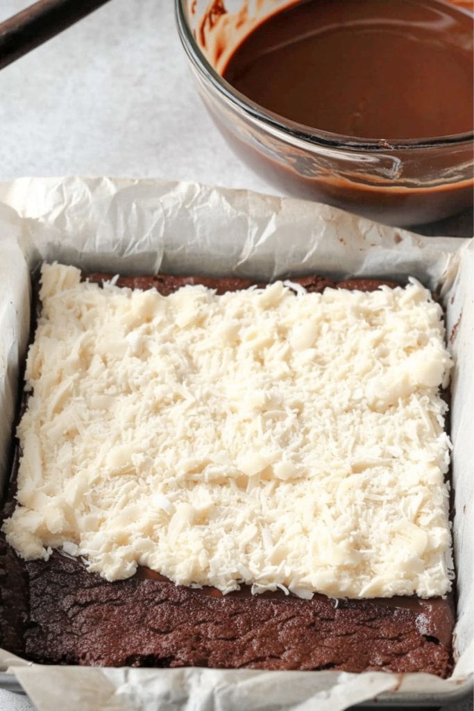 Brownies in a baking pan with parchment paper covered with coconut filling next to a glass bowl with chocolate ganache.