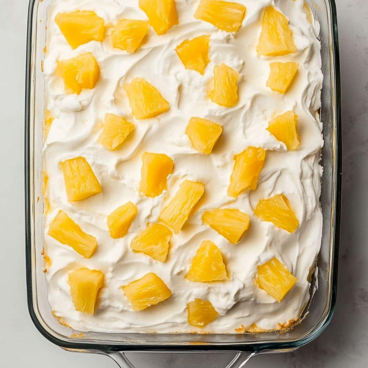 Whipped cream covered cake in a rectangular baking dish garnished with pineapple chunks on top.