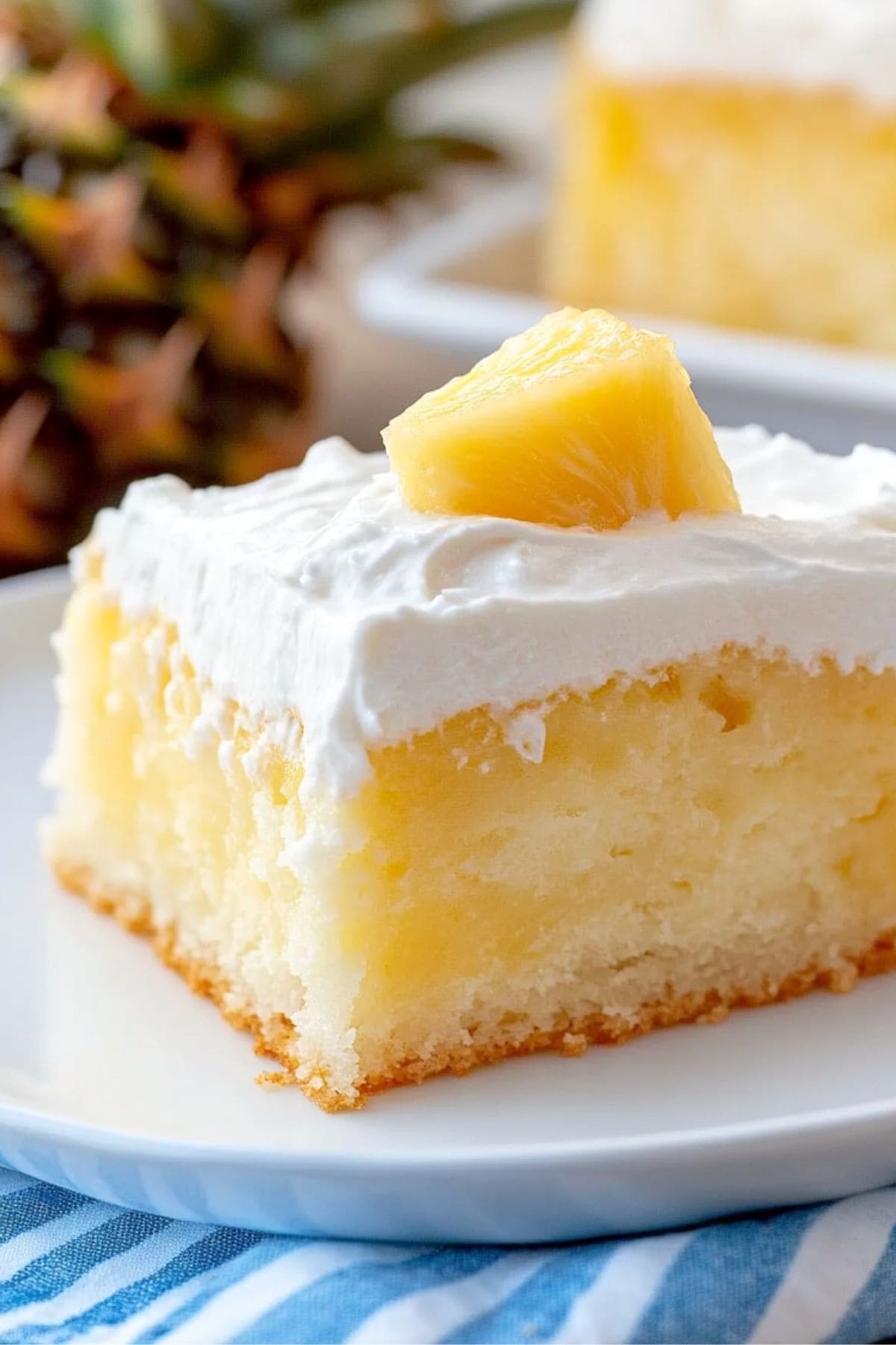 Pineapple poke cake square slice served in a white plate sitting on top of a white and blue stripped cloth.