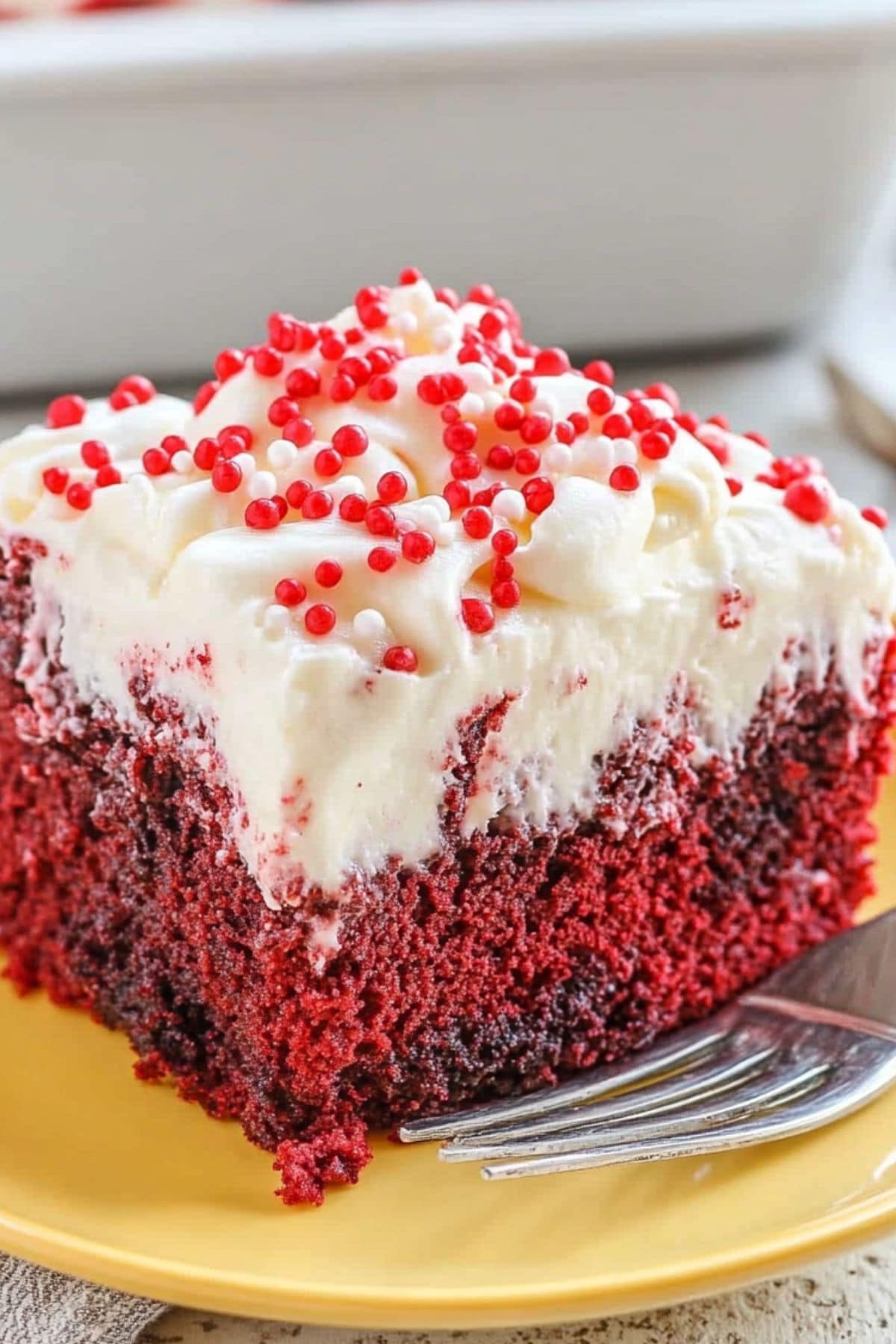 Slice of moist red velvet poke cake with whipped cream on top garnished with sprinkles served on a yellow plate.