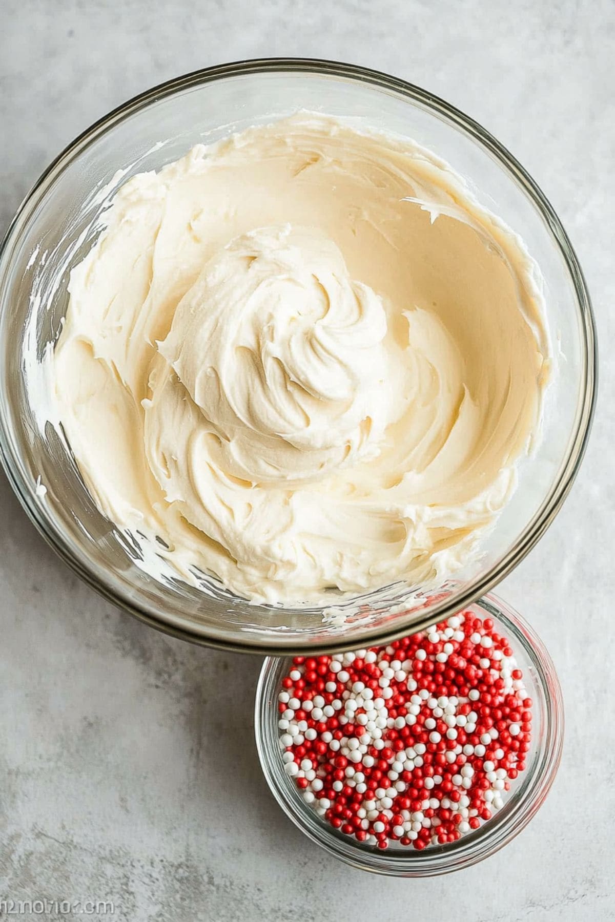 Frosting in  a glass bowl next to candy sprinkles in a glass bowl.