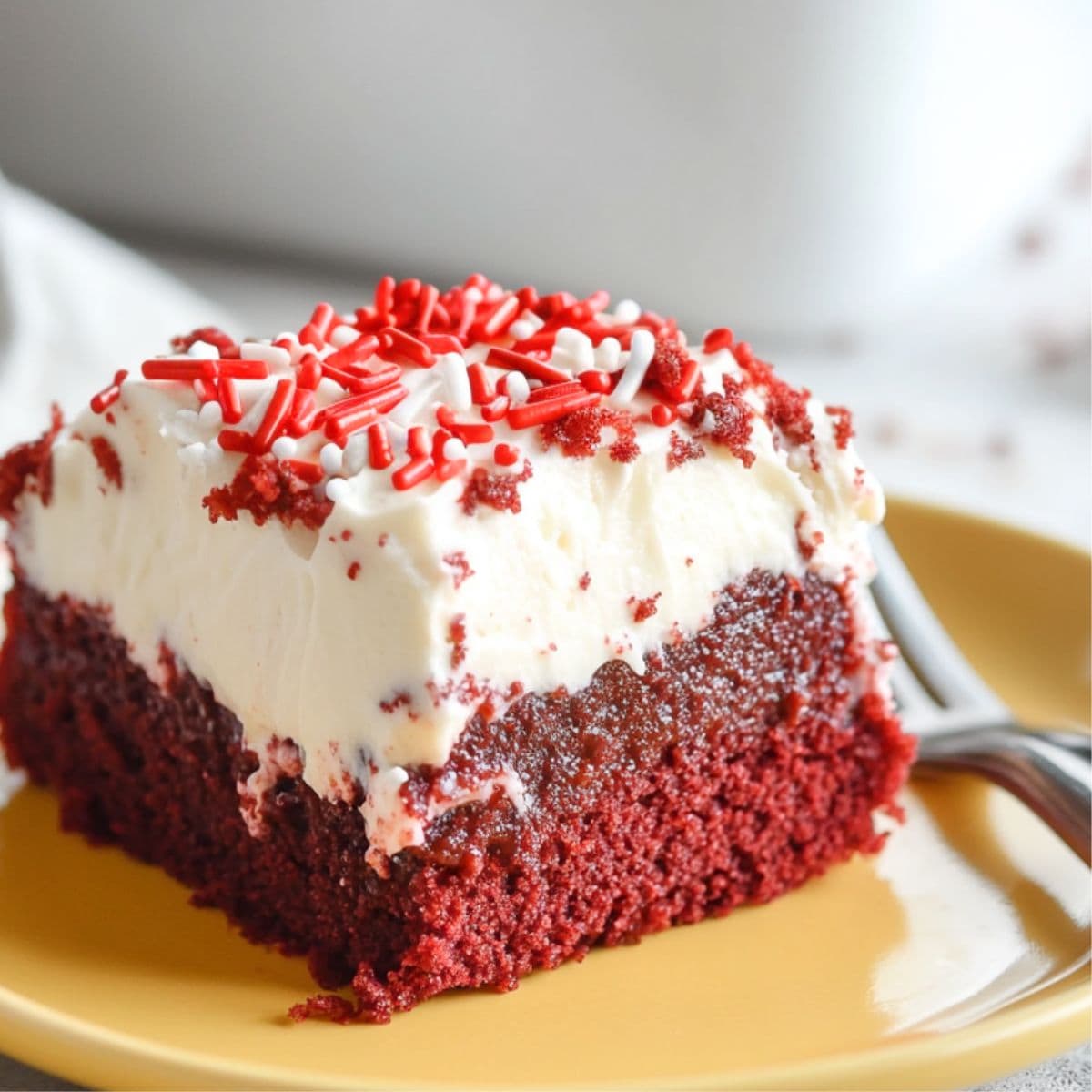 Square slice of red velvet poke cake with whipped cream on top garnished with candy sprinkles served in a yellow plate.