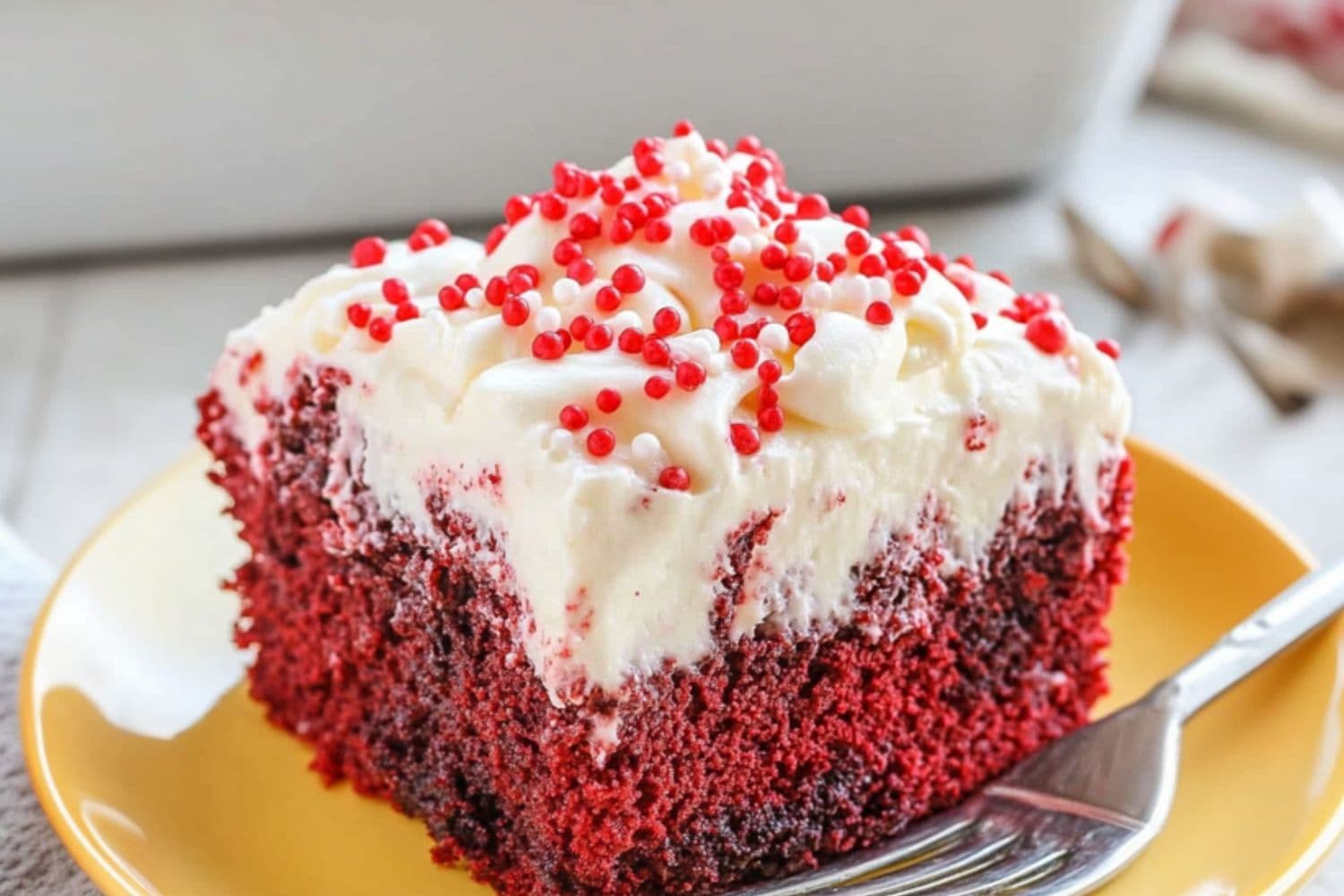 Square slice of red velvet cake covered with whipped cream garnished with candy sprinkles served on a yellow plate.