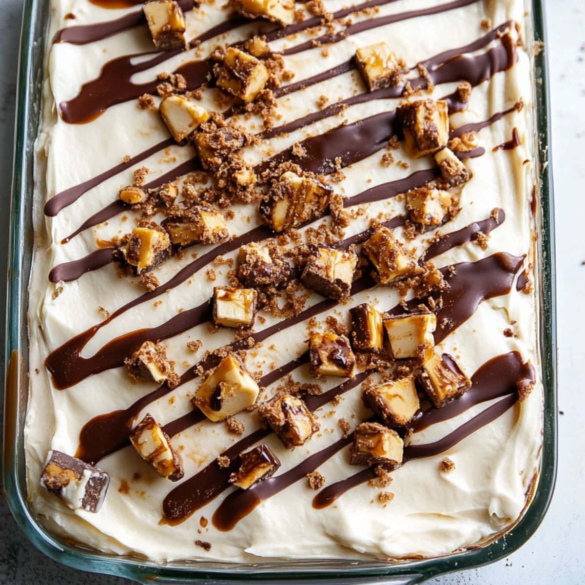 Snicker poke cake drizzled with chocolate syrup served in a glass dish.