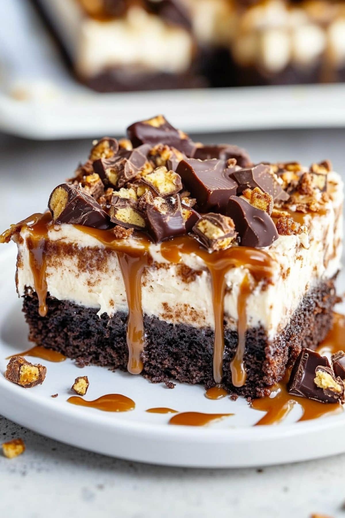 Square slice of chocolate poke cake with chopped snickers on top drizzled with caramel syrup.