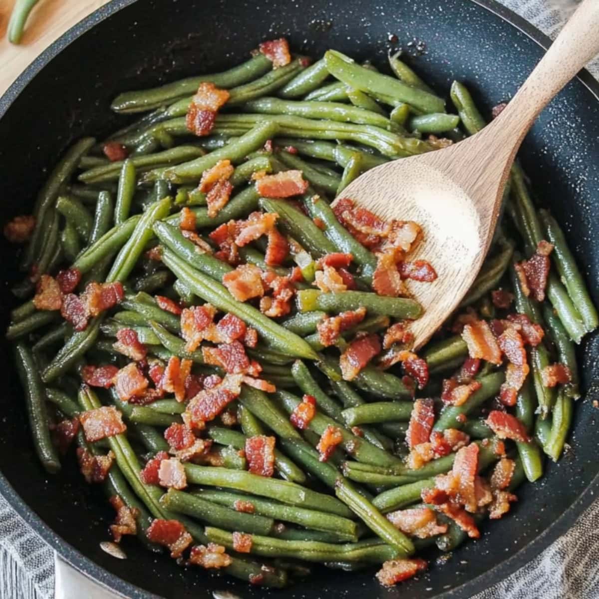 Wooden spoon tossing sauteed green beans with bacon cooked in a large pan. 