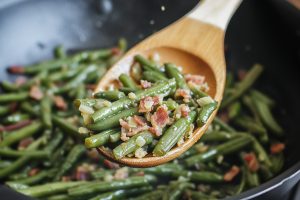 Sauteed bacon ang green beans tossed by a wooden spoon in a pan.