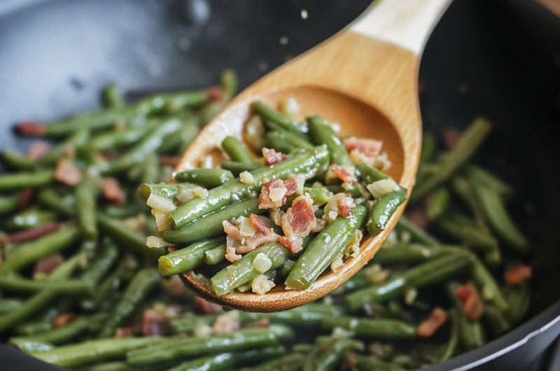 Sauteed bacon ang green beans tossed by a wooden spoon in a pan.