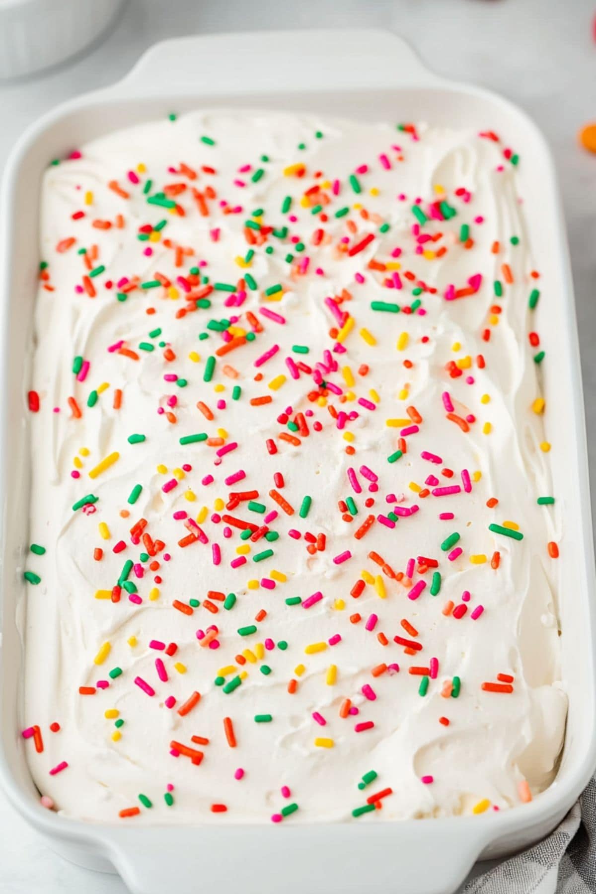 Vanilla cake in a rectangular baking dish covered with whipped cream and sprinkled with candy sprinkles.