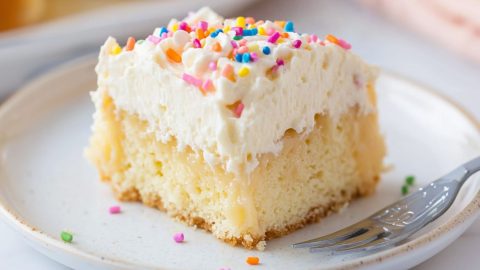 Vanilla poke cake slice with candy sprinkles on top served in a white plate.