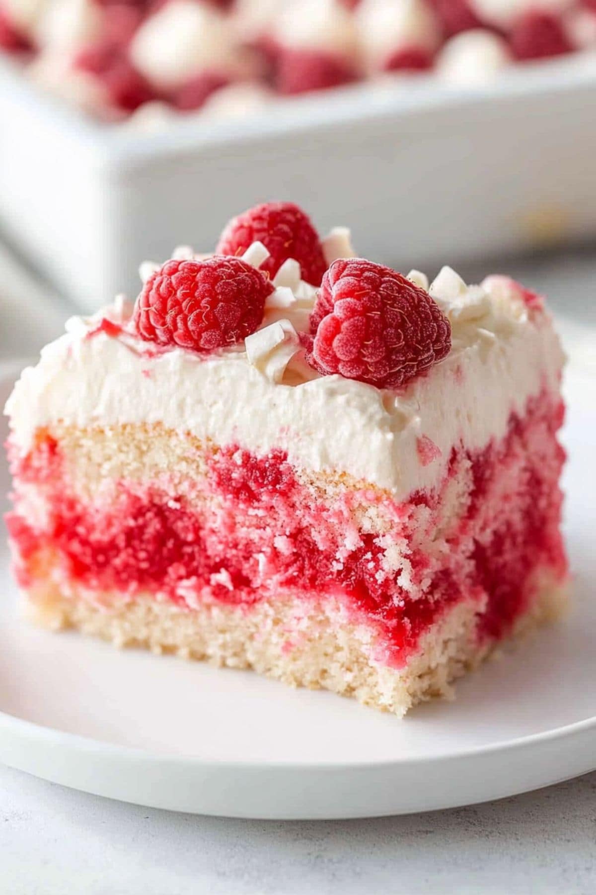 Poke cake with marbles of red Jello, topped with whipped cream and garnished with raspberries and white chocolate shavings.