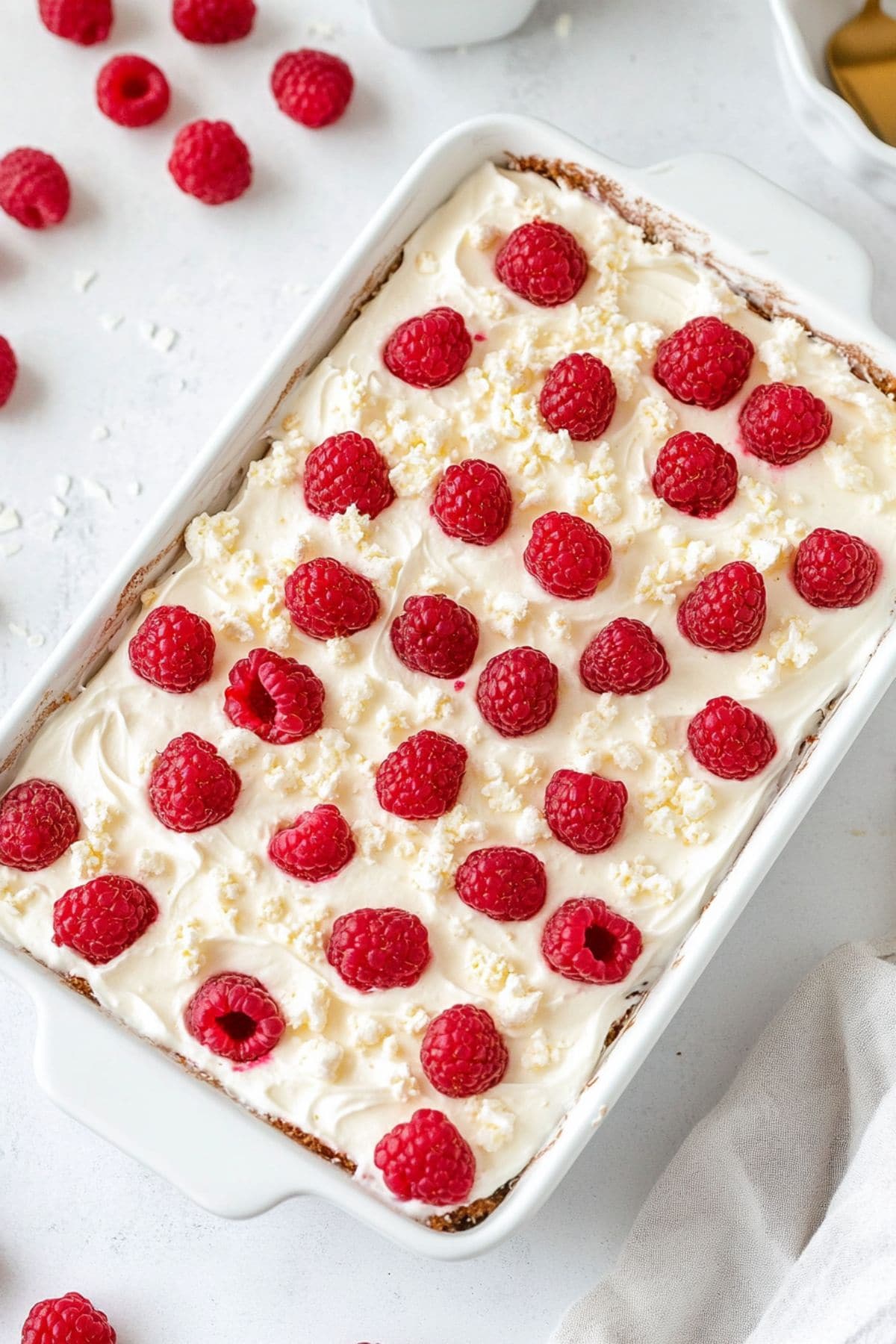 Cake baked in a rectangular white baking dish covered with whipped cream garnished with raspberries and white chocolate shavings on top.