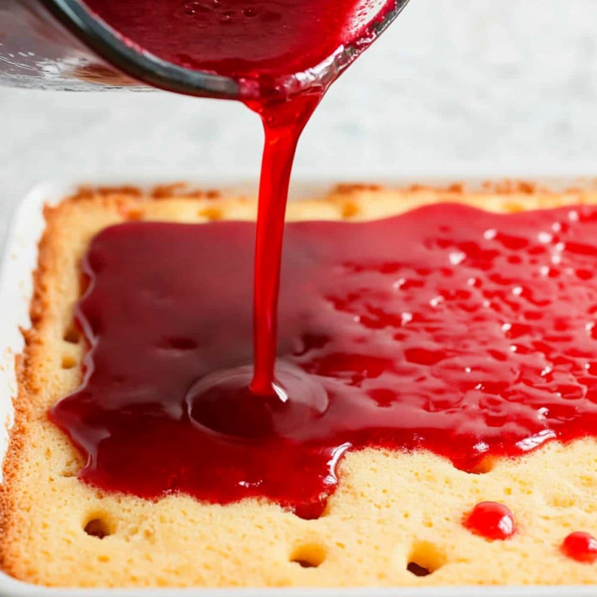 Red jello poured over vanilla cake with holes baked in rectangular white baking dish.