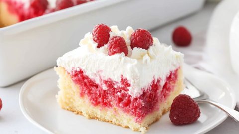 Serving slice of white chocolate raspberry poke cake in a white plate garnished with fresh raspberries and white chocolate shavings.