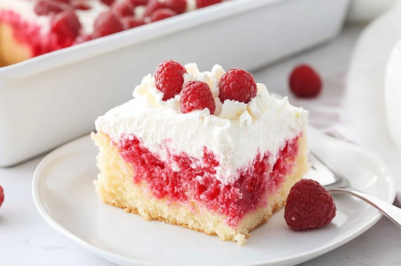 Serving slice of white chocolate raspberry poke cake in a white plate garnished with fresh raspberries and white chocolate shavings.