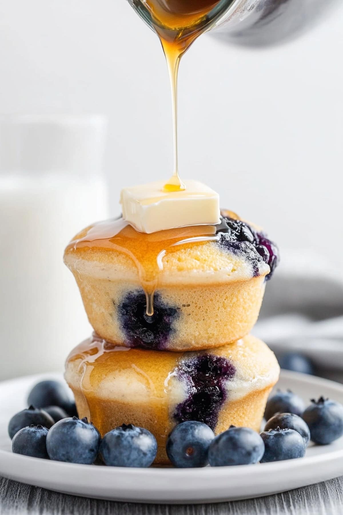 Two blueberry pancake muffins with a slice of butter on top drizzled with maple syrup served in a white plate. 