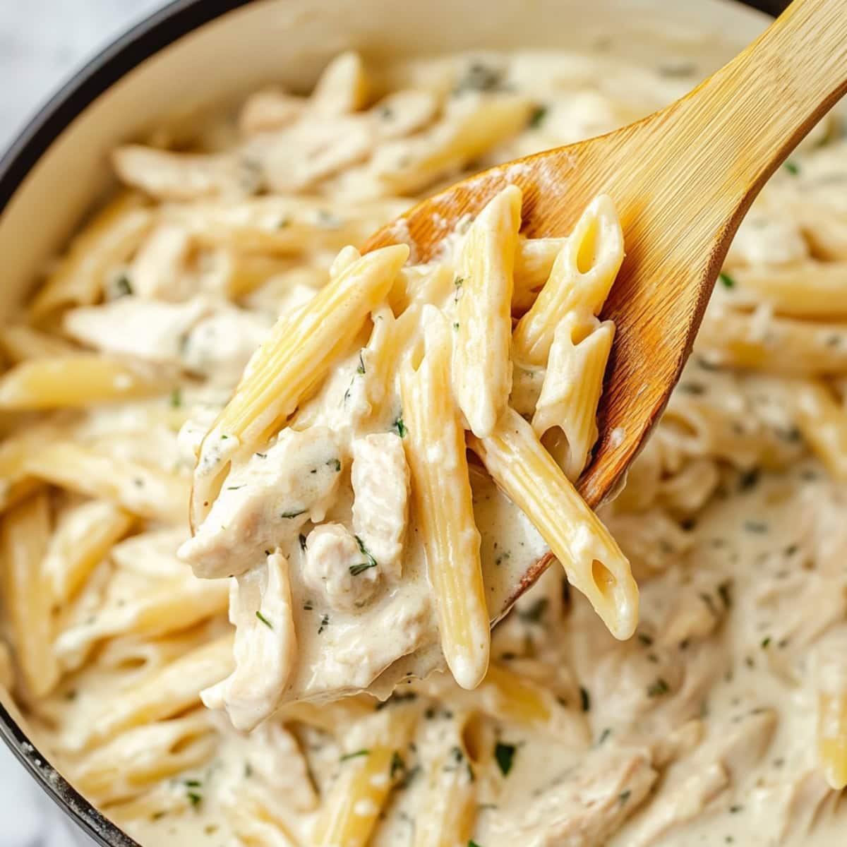 Wooden spoon scooping a serving of creamy chicken penne pasta from pot.