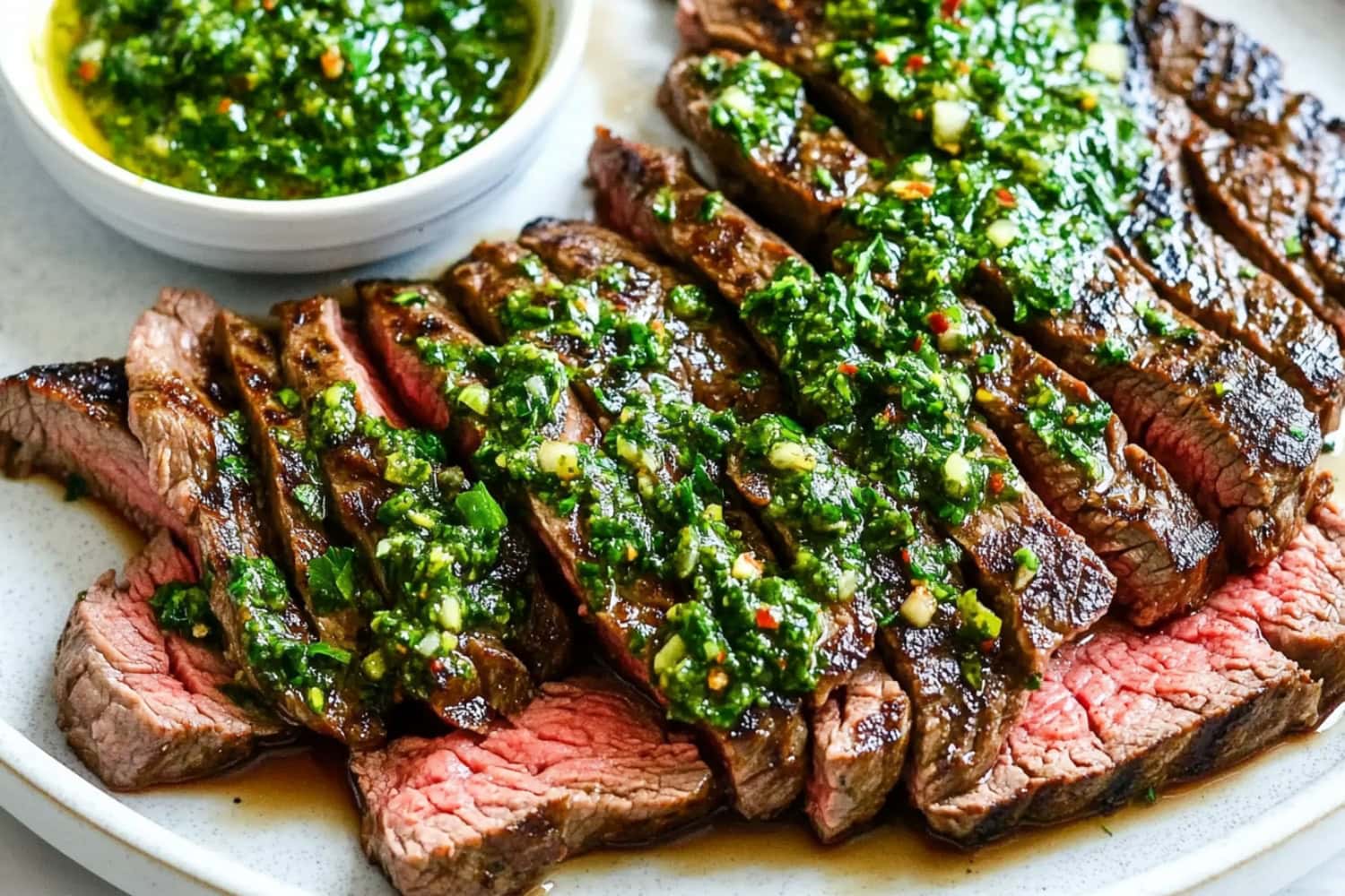 Sliced flank steak covered in a zesty chimichurri sauce, arranged on a white plate.