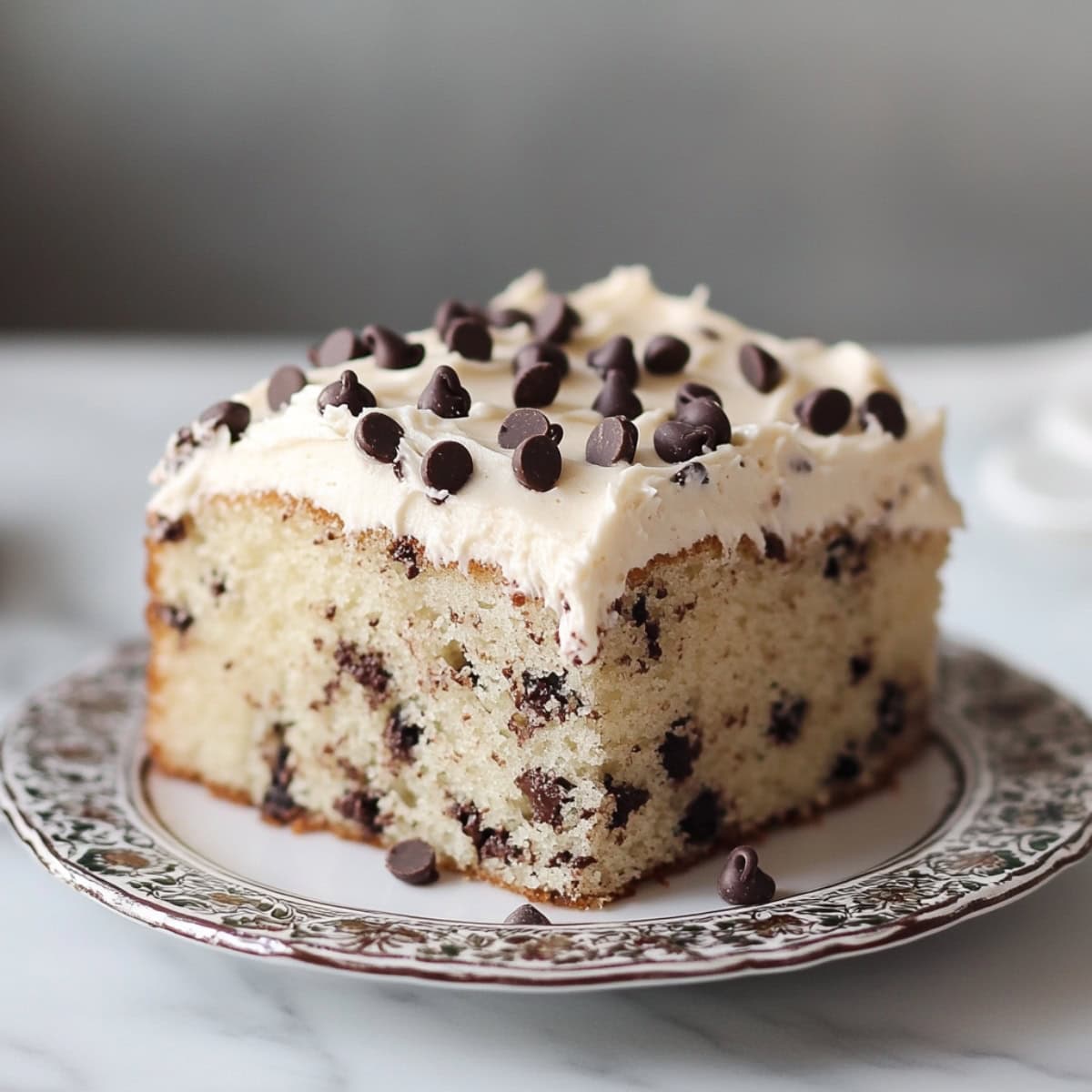 Square slice of chocolate chip sheet cake with white frosting and chocolate chips on top on a plate .