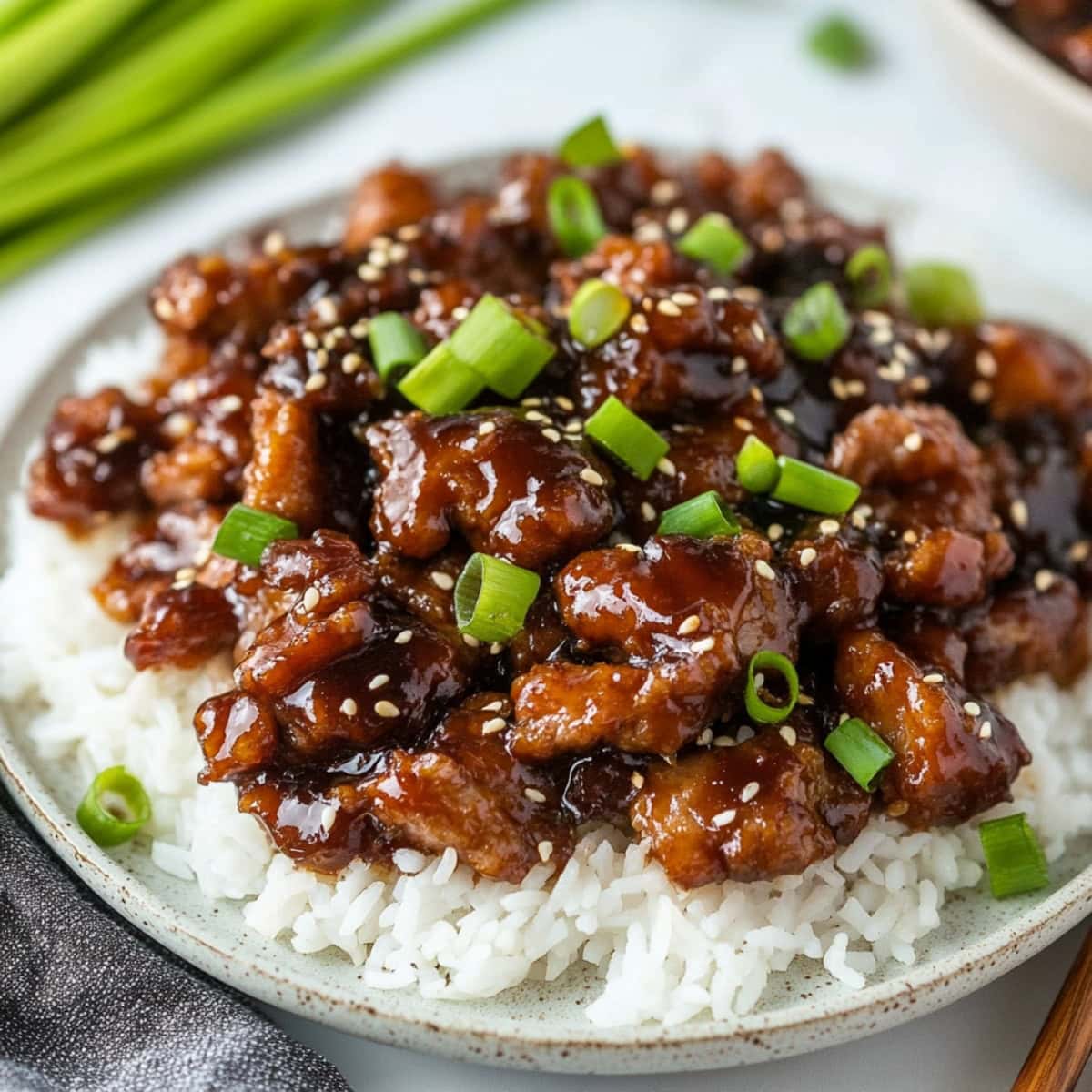 Mongolian pork with sauce garnished with chopped onions and sesame seeds served on top of white rice in a plate.