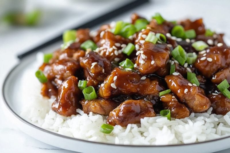 Mongolian pork served with white rice in a white plate garnished with sesame seeds and chopped onions.