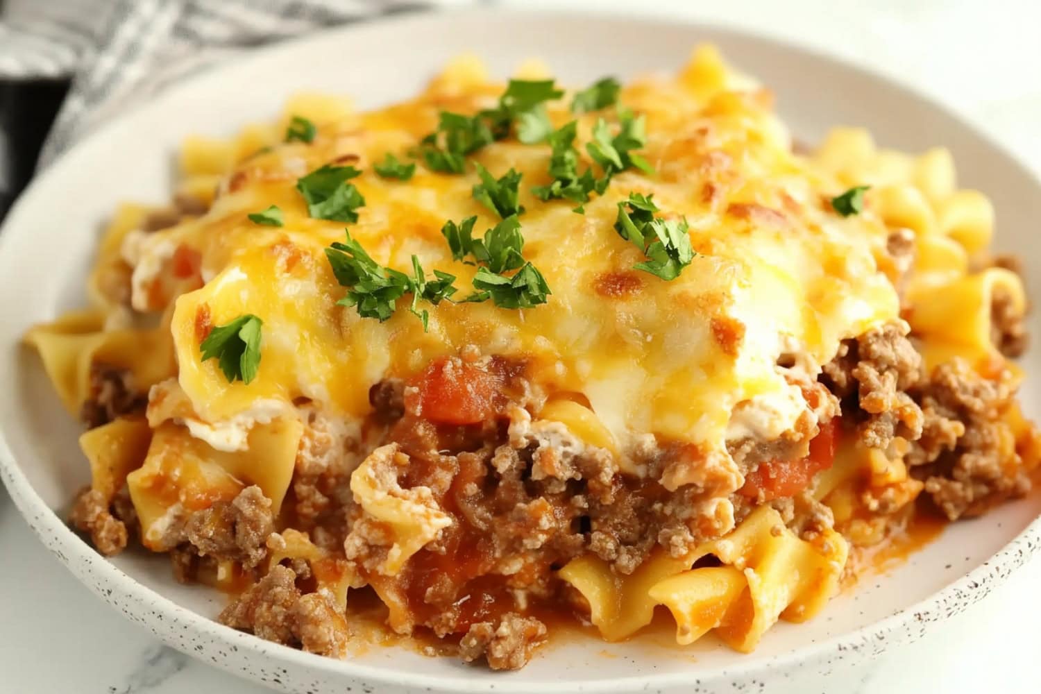 A portion of Beef Lombardi casserole on a plate, showcasing egg noodles and tomatoes, garnished with parsley.