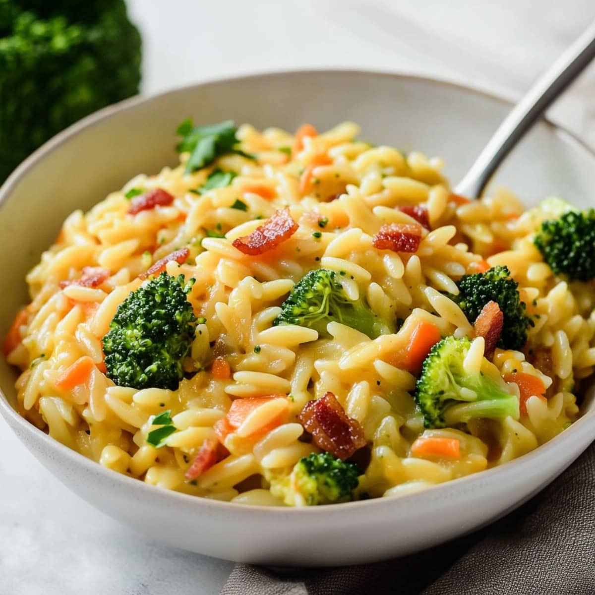 A close-up of a bowl of cheesy broccoli cheddar orzo with crispy bacon and shredded carrots.