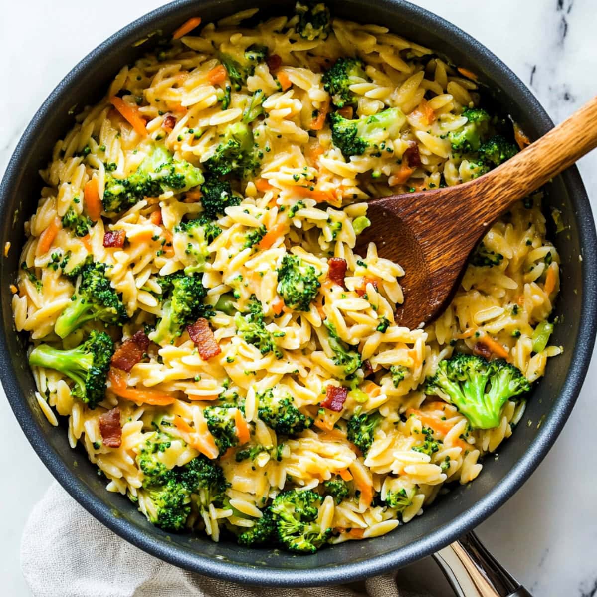 A saucepan of homemade broccoli cheddar orzo with bacon bits and shredded carrots.