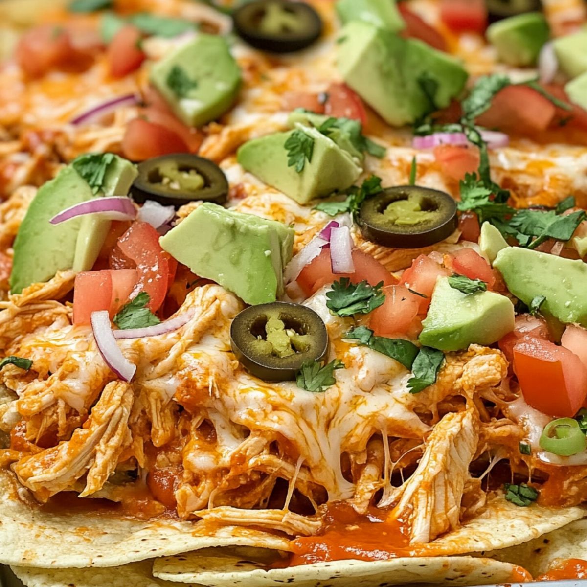 Buffalo chicken nachos in a sheet pan topped with melted cheese, garnished with chopped tomatoes, avocados and olives.