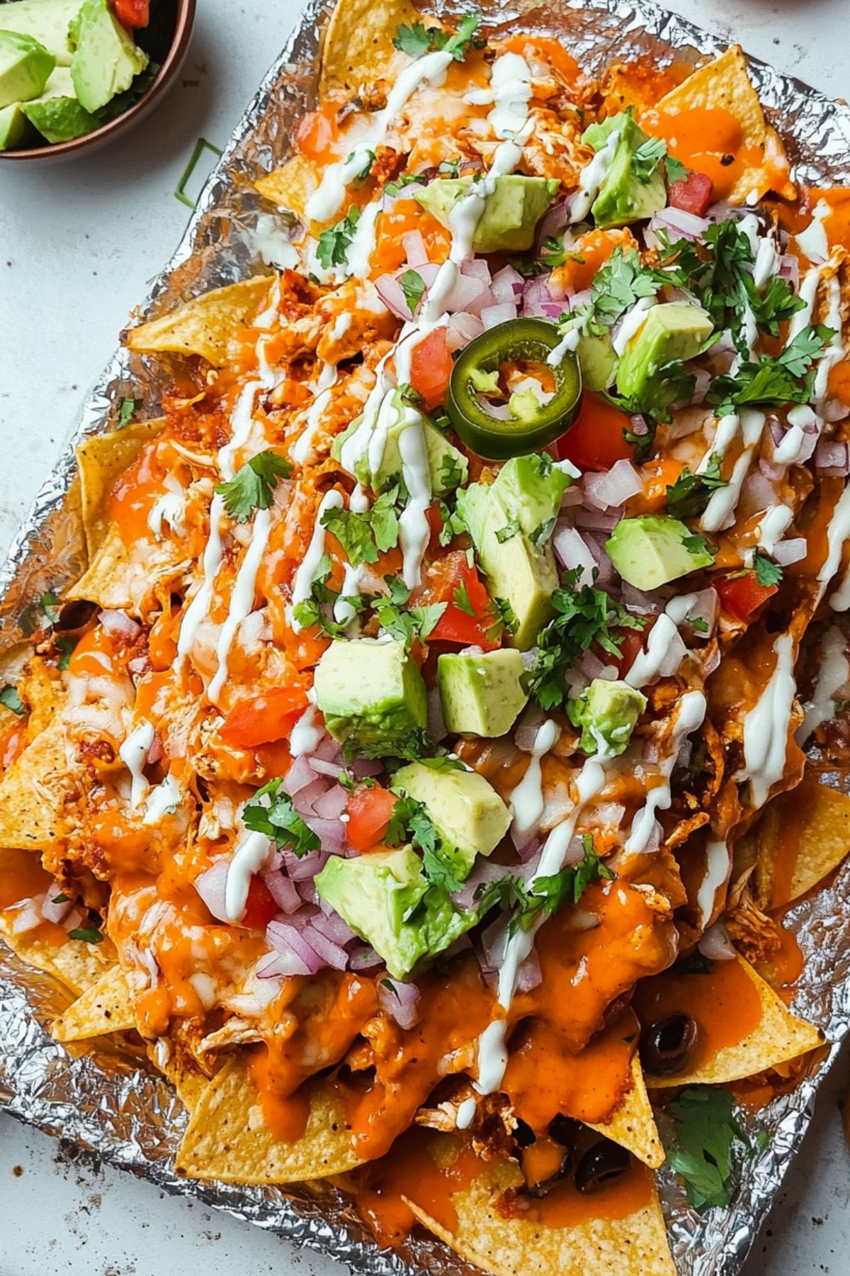 Bed of nachos in a sheet pan with aluminum foil covered with buffalo sauce and melted cheese drizzled with ranch dressing garnished with chopped avocado, tomatoes and pico de gallo.