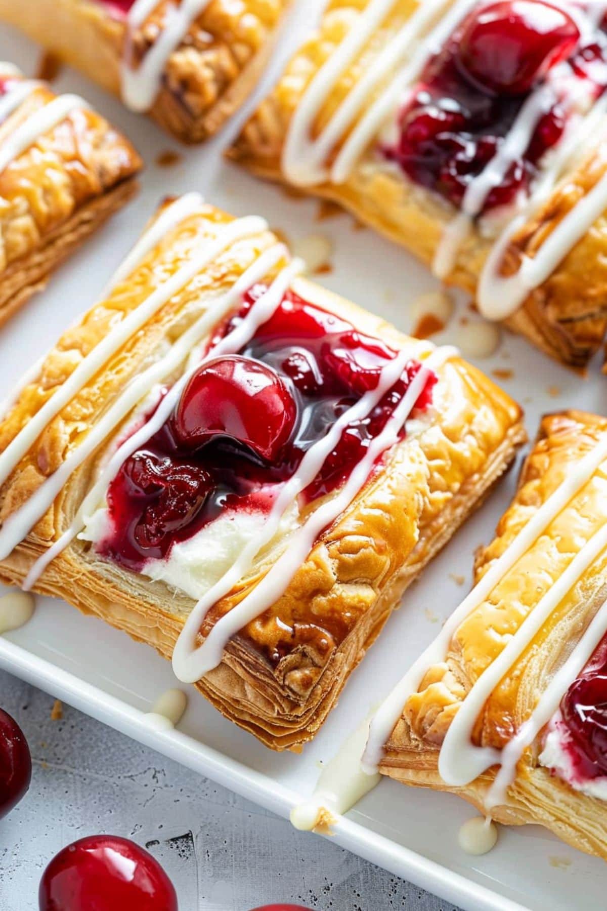 Bunch of puff pastry with cream cheese and cherry pie filling in the middle sitting on a white tray drizzled with glaze.