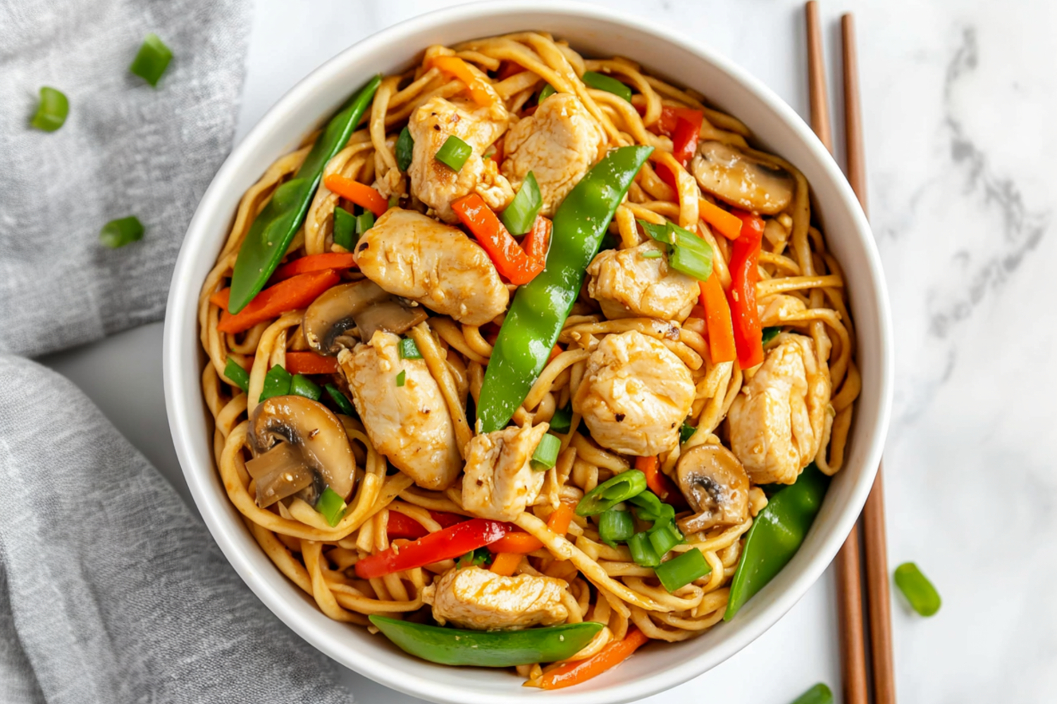 A skillet of chicken lo mein in a bowl with snow peas, carrots and bell peppers.