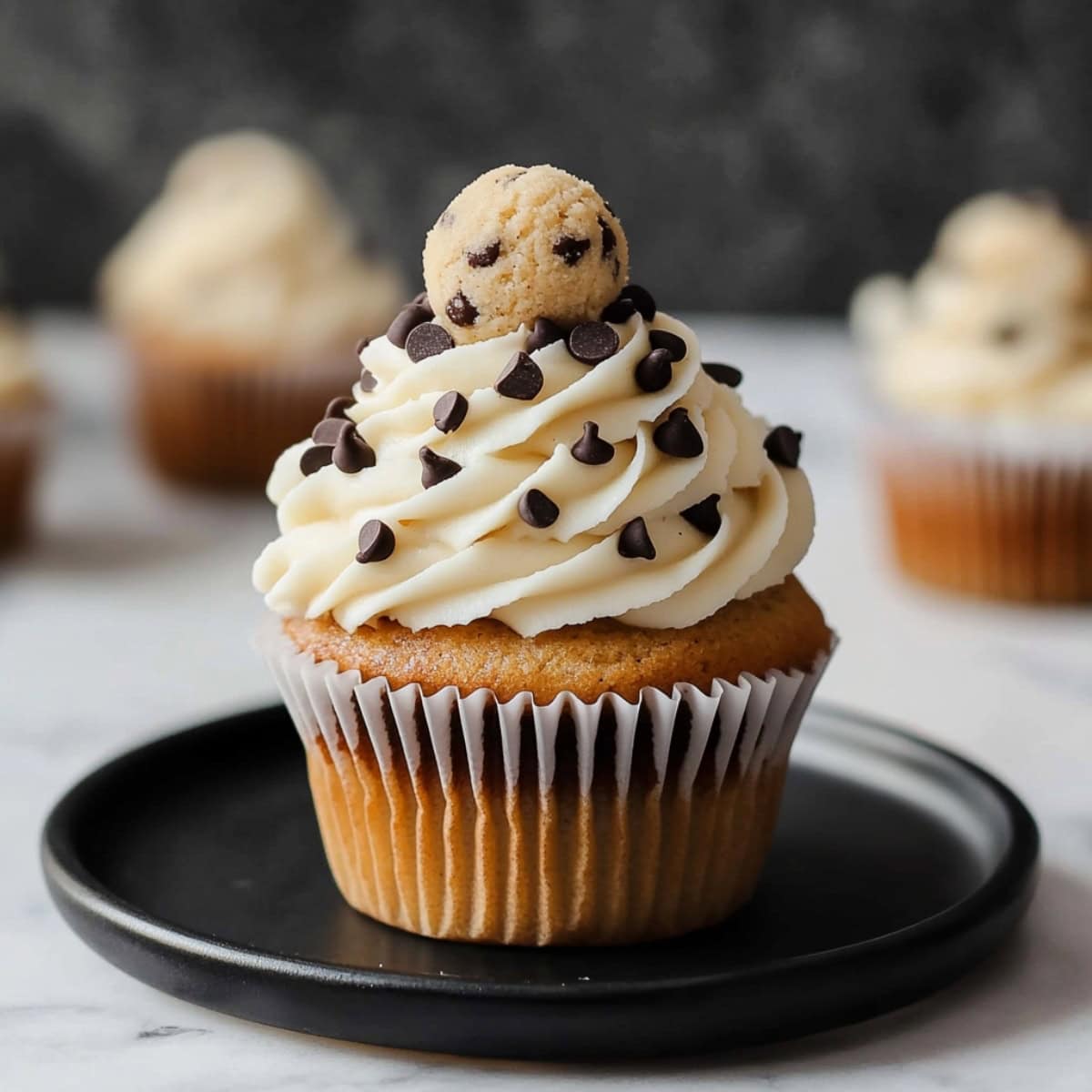 Close-up of a cookie dough cupcake with frosting and chocolate chips on a black plate.