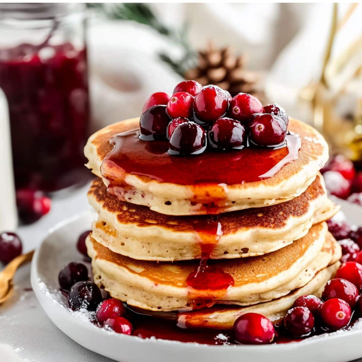 Pancakes served in a white plate drizzled with syrup, garnished with cranberries.