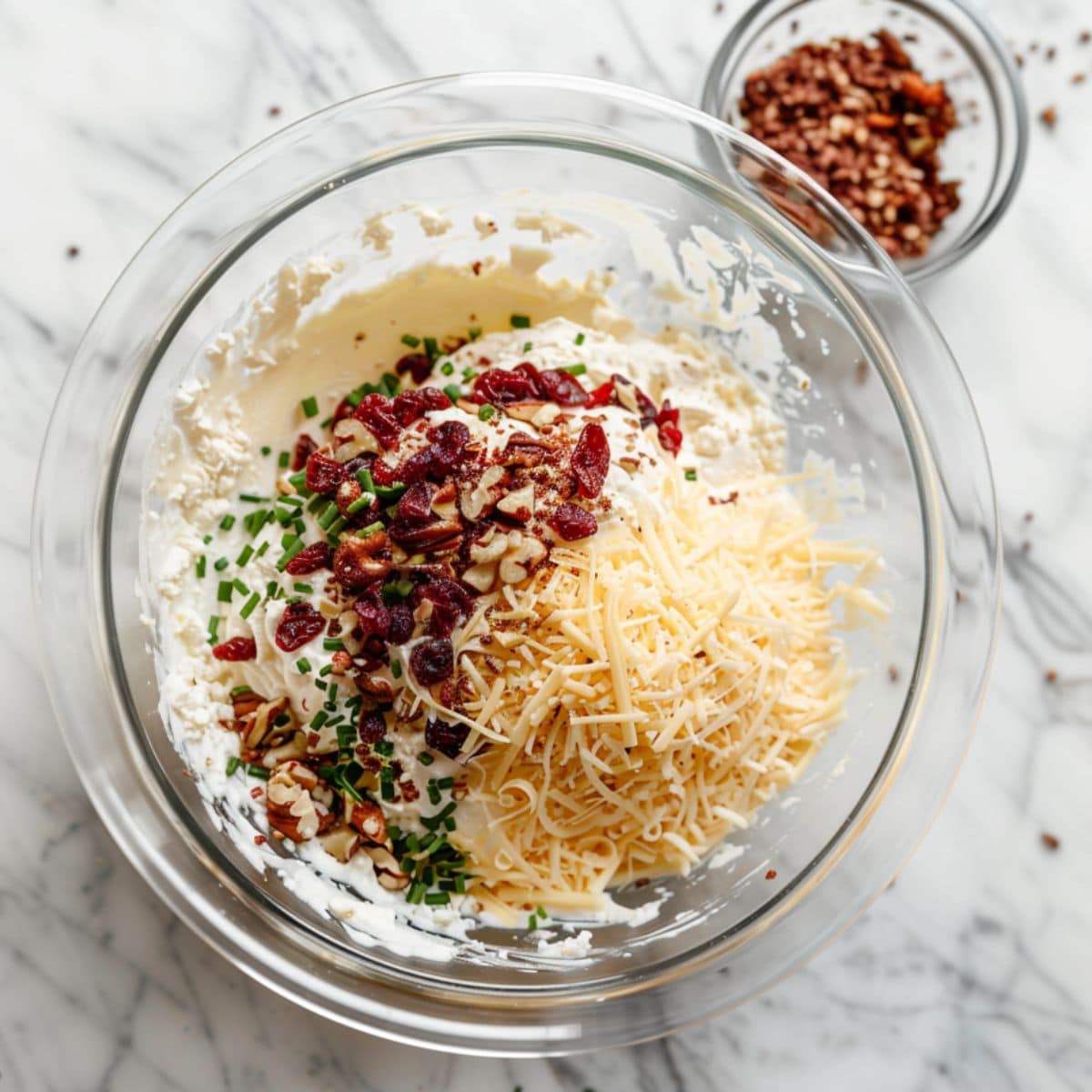 Cream cheese with grated cheese, cranberries, nuts and chives mixed in a glass mixing bowl.