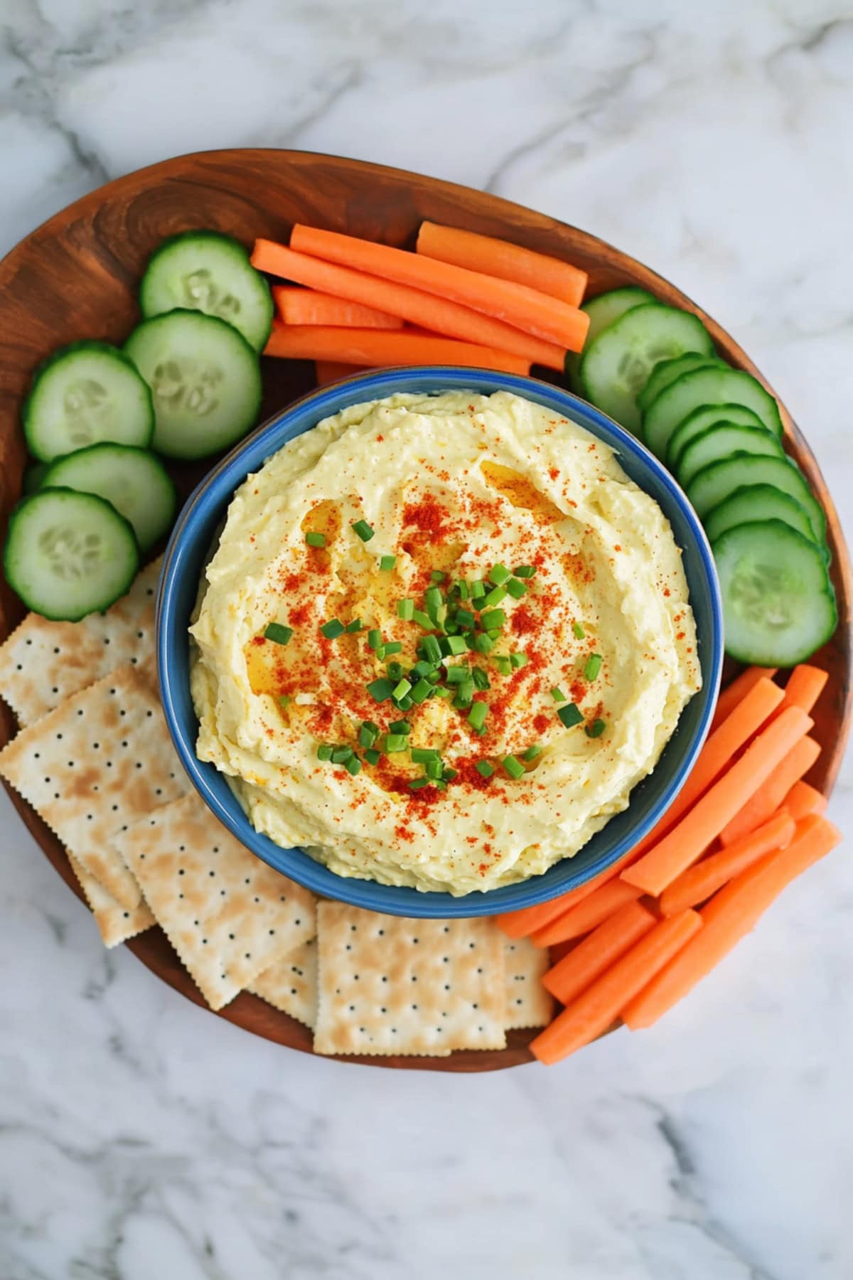 Creamy deviled egg dip with crackers, cucumbers and carrots on the side in a wooden plate.