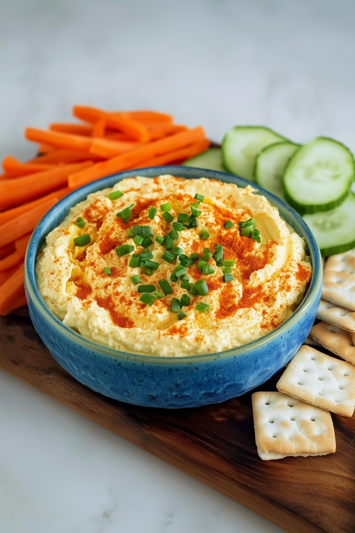 Deviled egg dip with paprika and chopped chives in a blue bowl, vegetables and crackers on the side.