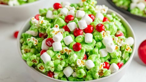 Popcorn coated and tossed in green coated melted marshmallows served in a white bowl garnished with red M&Ms and mini marshmallows.