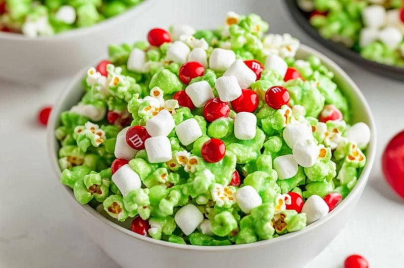 Popcorn coated and tossed in green coated melted marshmallows served in a white bowl garnished with red M&Ms and mini marshmallows.